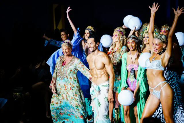 Luxeisle runway finale with people in colorful swimwear and robes, raised hands and white balloons